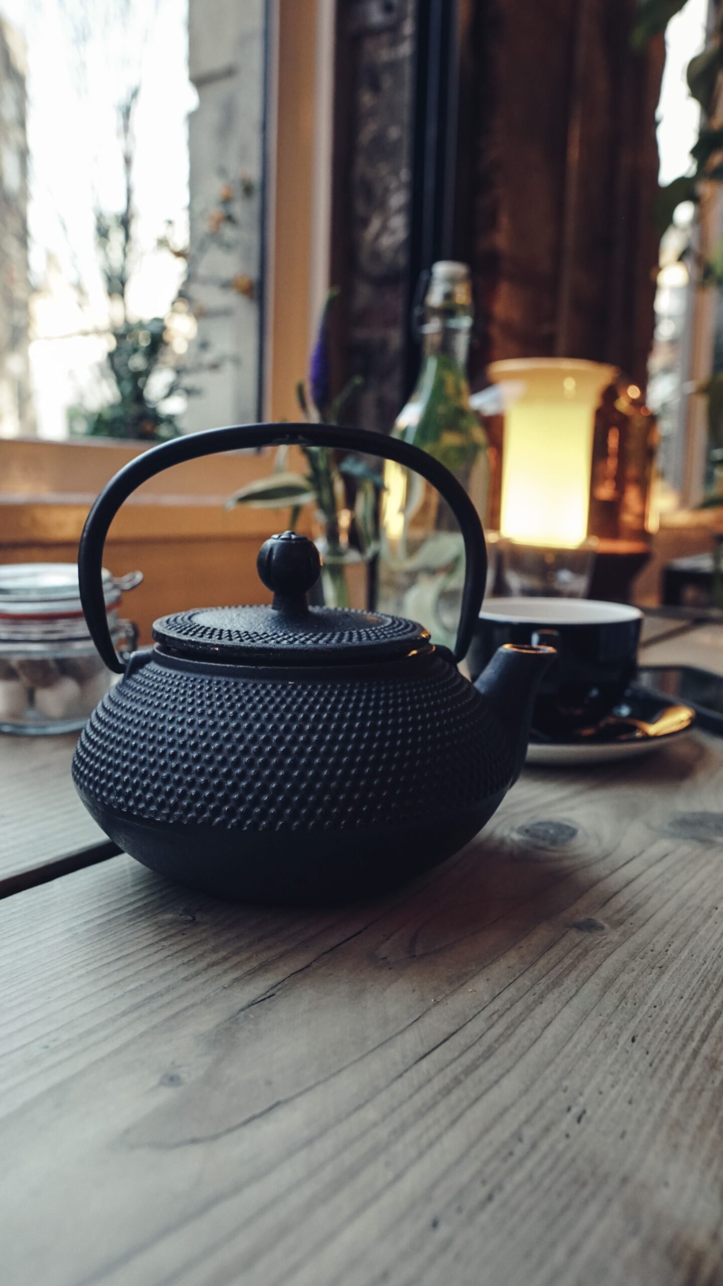 Black Japanese cast iron teapot with a textured finish displayed on a wooden table beside a cup and saucer, with warm lighting and plants in the background.